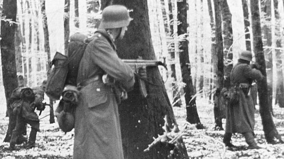 Deutsche Soldaten in einem verschneiten Wald. Im Vordergrund steht ein Soldat an einem Baum mit einem Sturmgewehr 44.