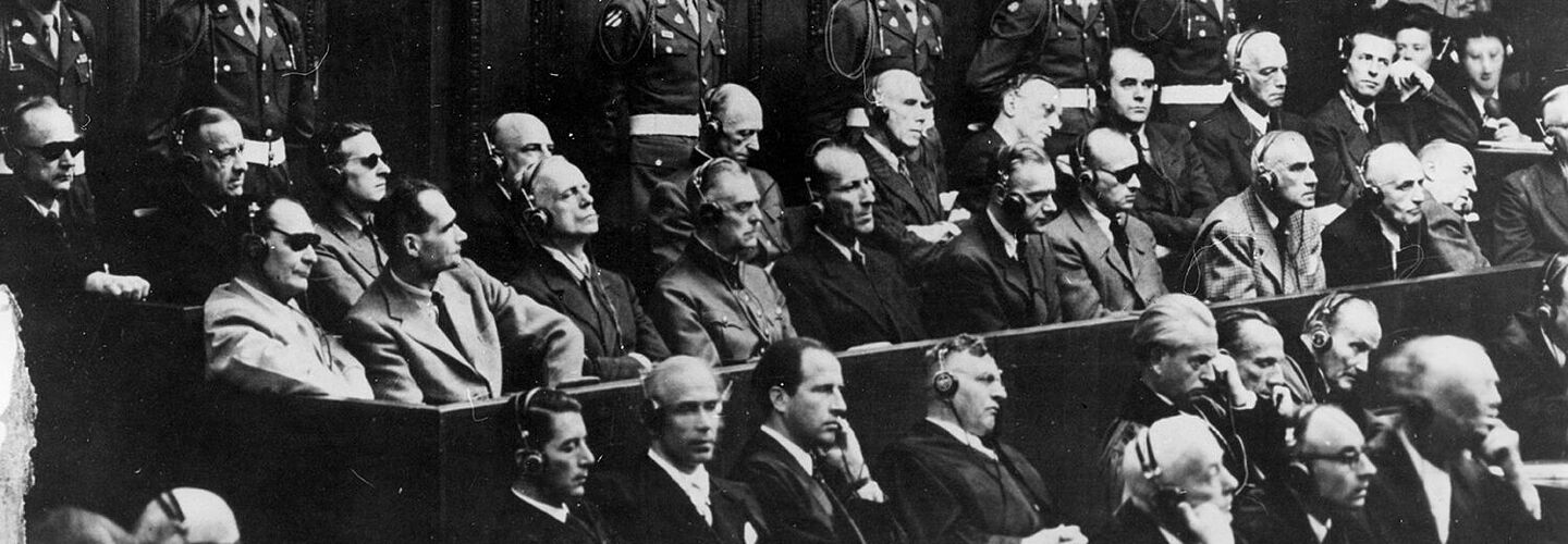 Blick auf die Anklagebank beim Nürnberger Prozess. Zu sehen sind die Hauptkriegsverbrecher Karl Dönitz, Erich Raeder, Baldur von Schirach, Fritz Sauckel, Alfred Jodl, Franz von Papen, Arthur Seyß-Inquart, Albert Speer, Konstantin von Neurath, Hans Fritsche, Hermann Göring, Rudolf Hess, Joachim von Ribbentrop, Wilhelm Keitel, Ernst Kaltenbrunner, Alfred Rosenberg, Hans Frank, Wilhelm Frick, Julius Streicher, Walter Funk und Hjalmar Schacht. Hinter den Angeklagten stehen amerikanische Militärpolizisten mit weißen Helmen.
