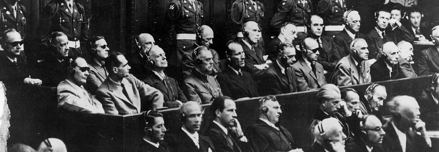 Blick auf die Anklagebank beim Nürnberger Prozess. Zu sehen sind die Hauptkriegsverbrecher Karl Dönitz, Erich Raeder, Baldur von Schirach, Fritz Sauckel, Alfred Jodl, Franz von Papen, Arthur Seyß-Inquart, Albert Speer, Konstantin von Neurath, Hans Fritsche, Hermann Göring, Rudolf Hess, Joachim von Ribbentrop, Wilhelm Keitel, Ernst Kaltenbrunner, Alfred Rosenberg, Hans Frank, Wilhelm Frick, Julius Streicher, Walter Funk und Hjalmar Schacht. Hinter den Angeklagten stehen amerikanische Militärpolizisten mit weißen Helmen.