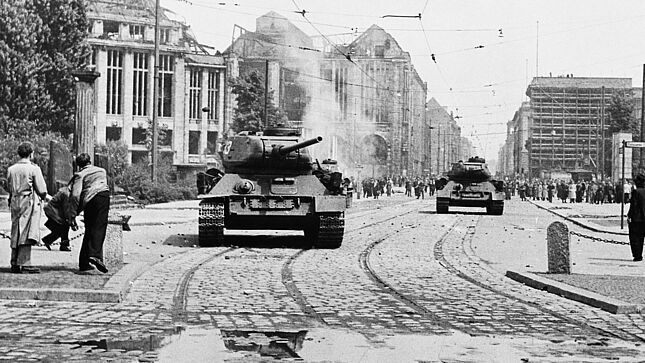 Das Schwarz-Weiß-Bild zeigt eine Straßenszene am 17. Juni 1953 in Berlin. Etwas versetzt befinden sich zwei Panzer auf der Straße. Im Vordergrund sind zwei junge Männer zu sehen, die gerade Steine auf die Panzer werfen.