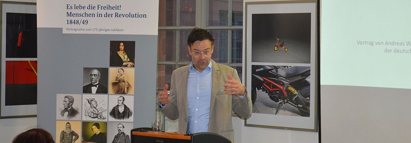 Andreas Weiß spricht in der Erinnerungsstätte Rastatt über den Revolutionär Daniel Heinrich Saul. Links neben ihm steht ein Aufsteller mit Bildern historischer Persönlichkeiten und dem Titel der Vortragsreihe „Es lebe die Freiheit! Menschen in der Revolution 1848/49“.