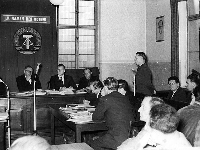 Mehrere Menschen sitzen in einem Gerichtssaal. Eine Person steht und spricht in ein Mikrofon. Im Hintergrund ist an der Wand das Staatswappen der DDR zu sehen, über dem steht "Im Namen des Volkes".
