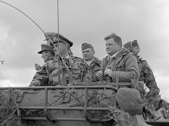 Verteidigungsminister Franz Josef Strauß beobachtet ein Manöver der Bundeswehr. Er steht zusammen mit fünf Soldaten auf einem gepanzerten Fahrzeug.