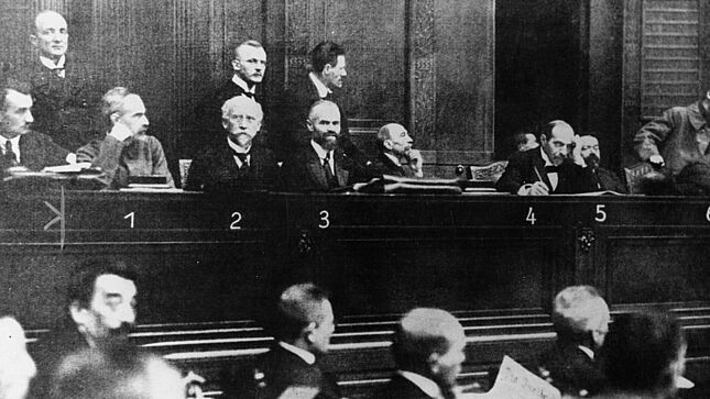 Schwarz-Weiß-Aufnahme eines mit sechs Herren in Anzügen besetzten Podiums mit einer Nummerierung in der Bildmitte und einer Legende mit Namen am unteren Bildrand