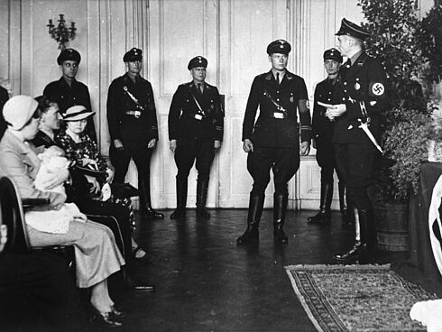 Schwarz-Weiß-Fotografie mit Teilnehmern einer Taufgesellschaft, darunter festlich mit Hüten gekleidete Frauen, davon eine mit einem Baby im Arm, sowie mehrere uniformierte Männer mit Hakenkreuzarmbinden.