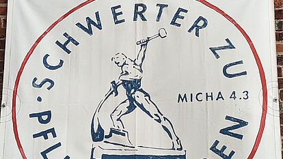 Eine Fahne mit dem Text: Schwerter zu Pflugscharen. In der Mitte des Bildes ein Mann, der aus einem Schwert ein Pflugteil schmiedet