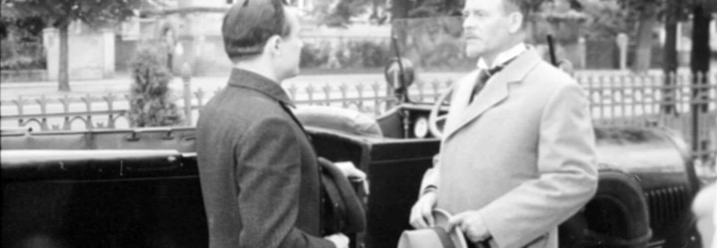 Das Bild zeigt eine Szene aus dem Film 'Mord an Rathenau' von 1961. Zwei Männer stehen sich vor einem Auto gegenüber und unterhalten sich.