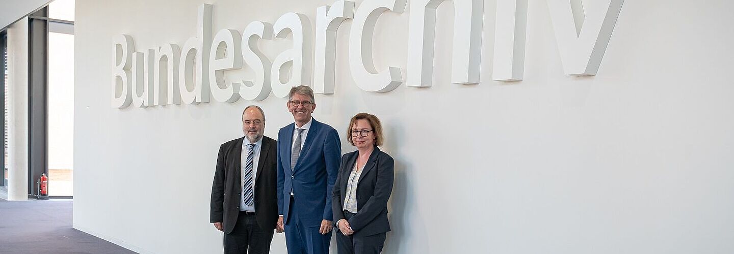 Michael Hollmann, Wolfram Weimer und Andrea Hänger stehen im Foyer des Bundesarchiv-Standorts Berlin-Lichterfelde vor dem Schriftzug „Bundesarchiv“.