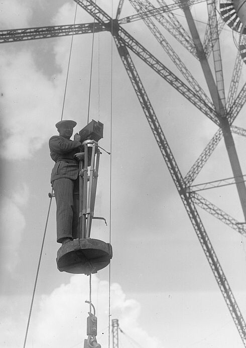 Schwarz-Weiß-Foto mit einem Mann, der auf einem kleinen Podest stehend aus der Luft Filmaufnahmen mit einer Standkamera macht