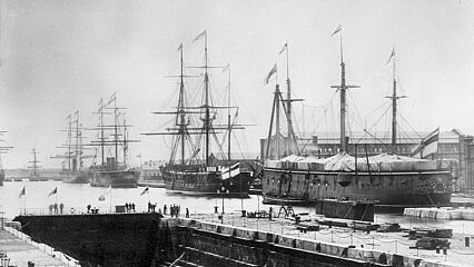 Mehrere Schiffe liegen in einem Hafen, auf dessen Kai Menschen stehen.