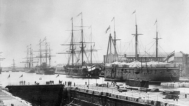 Mehrere Schiffe liegen in einem Hafen, auf dessen Kai Menschen stehen.