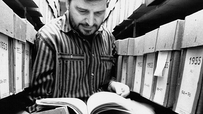 Ein Mann steht zwischen Aktenregalen und blättert in einer Stasi-Akte.