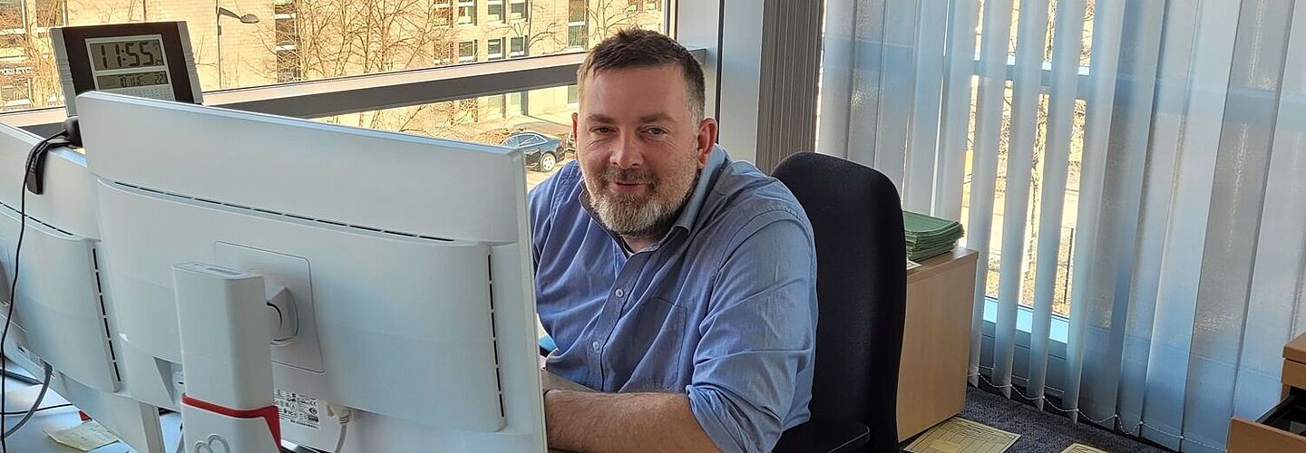 Robert-Christian Balsam sitzt an seinem Arbeitsplatz vor einem Monitor und blickt lächelnd in die Kamera.