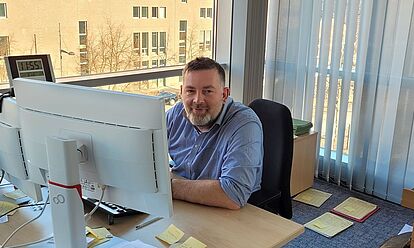 Robert-Christian Balsam sitzt an seinem Arbeitsplatz vor einem Monitor und blickt lächelnd in die Kamera.