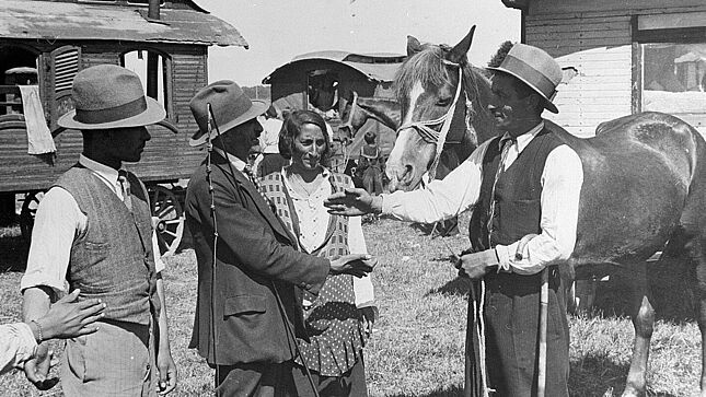 Schwarz-Weiß-Aufnahme mit drei Männern und einer Frau bei der Übergabe eines Pferdes sowie mit mehreren Wohnwagen im Hintergrund