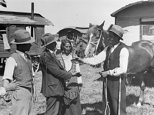 Schwarz-Weiß-Aufnahme mit drei Männern und einer Frau bei der Übergabe eines Pferdes sowie mit mehreren Wohnwagen im Hintergrund