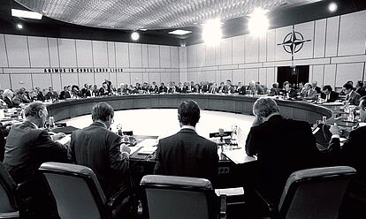 Die Außenminister der NATO-Staaten in einem großen Konferenzraum während einer Sondersitzung des NATO-Rats am 10. September 1990. An der Wand im Hintergrund steht das Motto der NATO „Animus in Consulendo Liber“ (Ein freier Geist in der Diskussion).