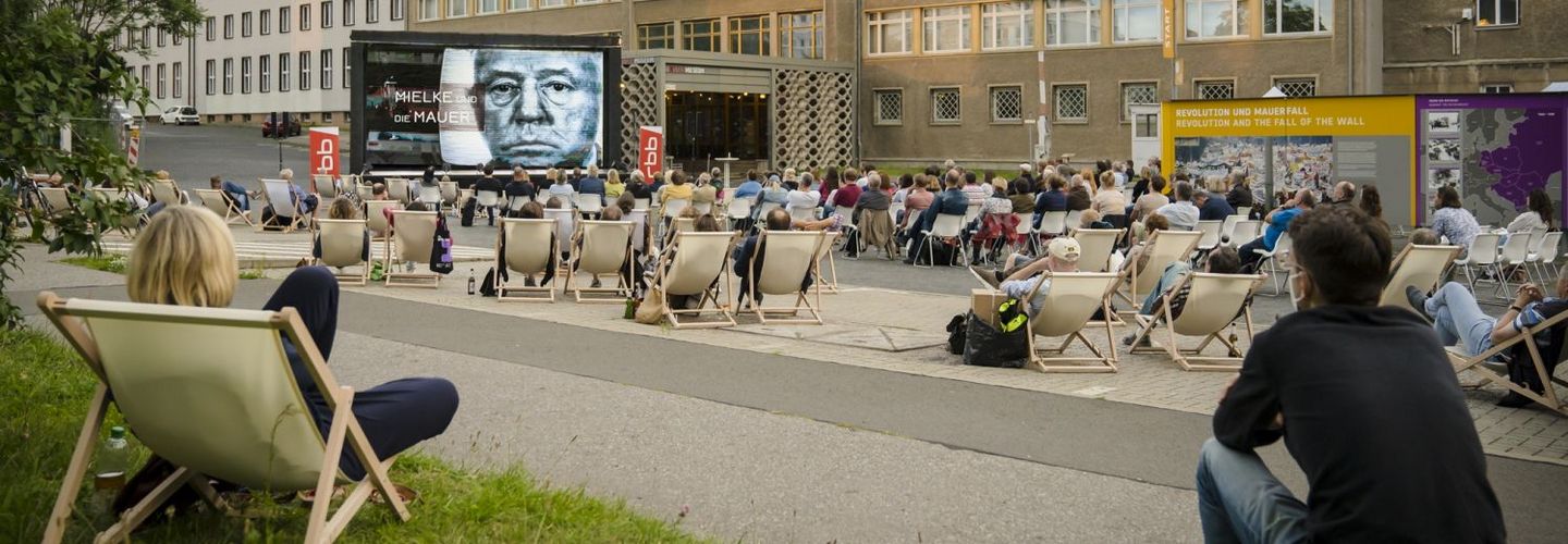 Vor Haus 1 steht die Kinoleinwand, auf die zahlreiche Zuschauer blicken. Auf der Leinwand ist das Gesicht Erich Mielkes zu erkennen.