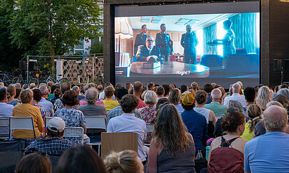 Menschen sitzen vor einer großen Leinwand und sehen den Spielfilm 'Leander Haußmanns Stasikomödie'.