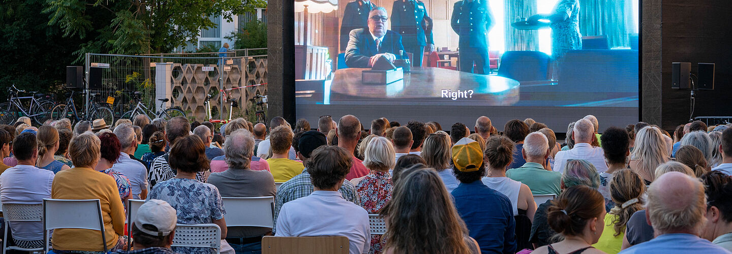 Menschen sitzen vor einer großen Leinwand und sehen den Spielfilm 'Leander Haußmanns Stasikomödie'.