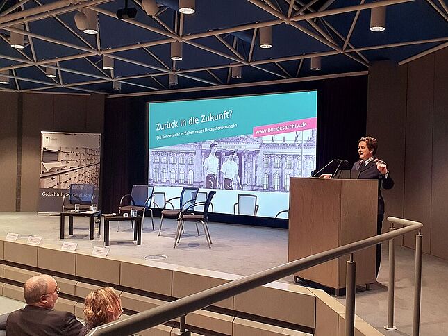 Nicole Schilling bei einer Veranstaltung zum Thema Bundeswehr Eine Frau in Uniform der Bundeswehr steht an einem Pult auf einer Bühne und spricht ins Mikrofon. Im Hintergrund wird ein Bild projiziert mit den Worten „Zurück in die Zukunft? Die Bundeswehr in Zeiten neuer Herausforderungen“.