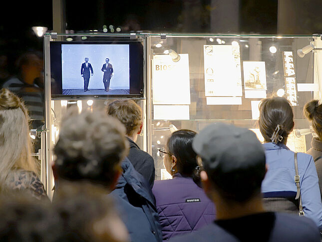 Eine mit Dokumenten gefüllte Vitrine ist zu sehen. Davor steht eine Gruppe von Menschen, die in Richtung der Vitrine schaut. Links an der Vitrine ist ein kleiner Bildschirm zu sehen, auf dem ein Film abgespielt wird.