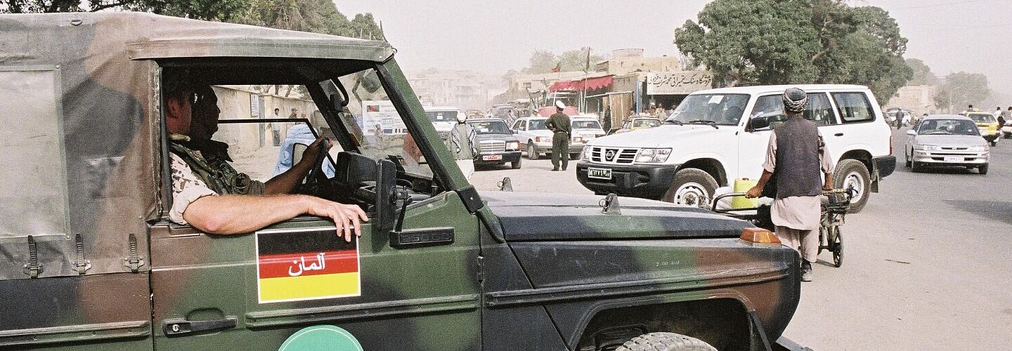 Ein Militärfahrzeug, auf das die deutsche Flagge gemalt ist, steht auf einer Straße in Afghanistan. Im Hintergrund sind Menschen und weitere Fahrzeuge zu sehen.