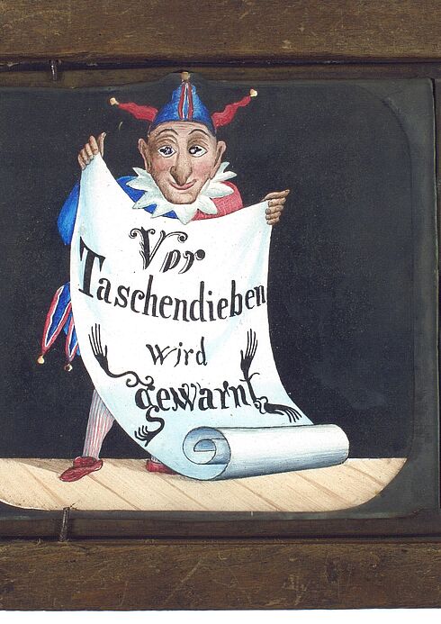 brauner Holzkasten mit eingefasster Glasscheibe, auf die ein Narr gezeichnet ist, der eine Papierrolle mit der Aufschrift "Vor Taschendieben wird gewarnt" in den Händen hält