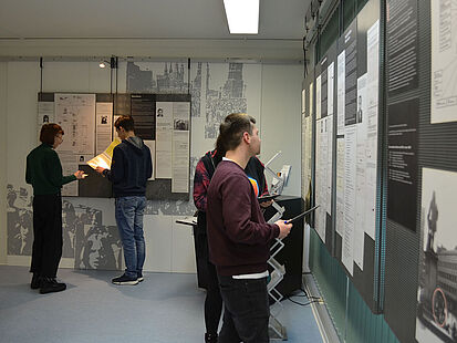 Vier junge Menschen stehen in einem Raum und sehen sich Ausstellungstafeln an.