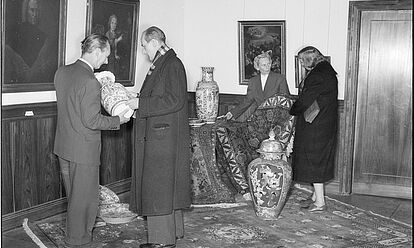 Innenraum mit drei großformatigen, alten Gemälden an der Wand. Davor 4 Personen, die vier große Porzellanvasen begutachten. Es handelt sich augenscheinlich um eine Verkäuferin, die eine Kundin im Pelzmantel berät und einen Verkäufer, der einem Käufer in langem Mantel eine Vase präsentiert. Auf dem Boden liegt ein wertvoller Teppich.