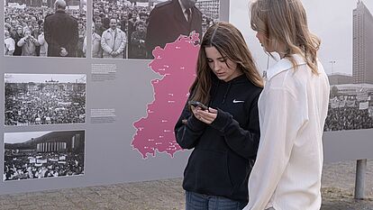 Zwei Schülerinnen stehen in einer Open Air Ausstellung und schauen auf ihre Handys.