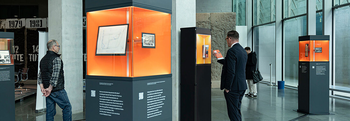Ausstellungsstelen verteilt in einem Raum mit Glasfront, Menschen betrachten die ausgestellten Objekte