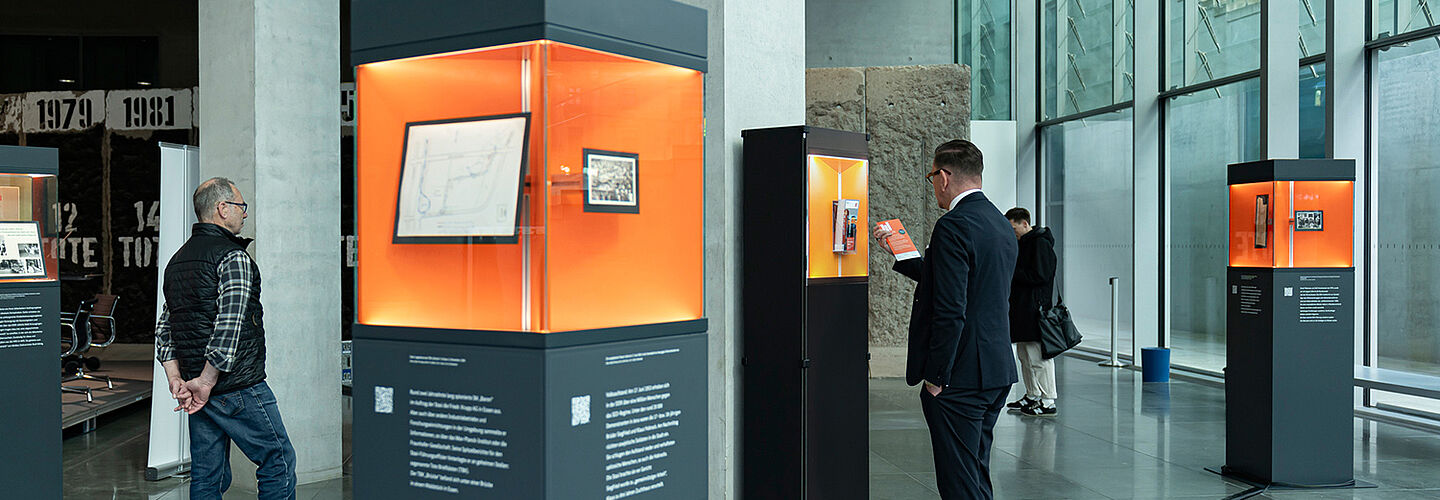 Ausstellungsstelen verteilt in einem Raum mit Glasfront, Menschen betrachten die ausgestellten Objekte