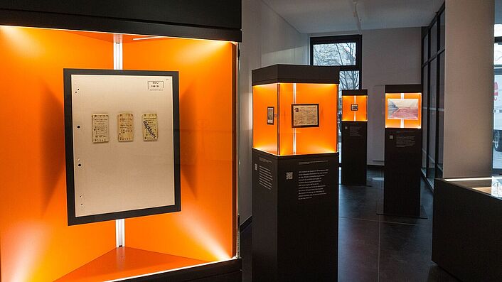 Blick in die Wanderausstellung. Zu sehen sind mehrere Stelen mit Glasvitrinen, in denen sich Dokumente, Fotos und Objekte befinden.