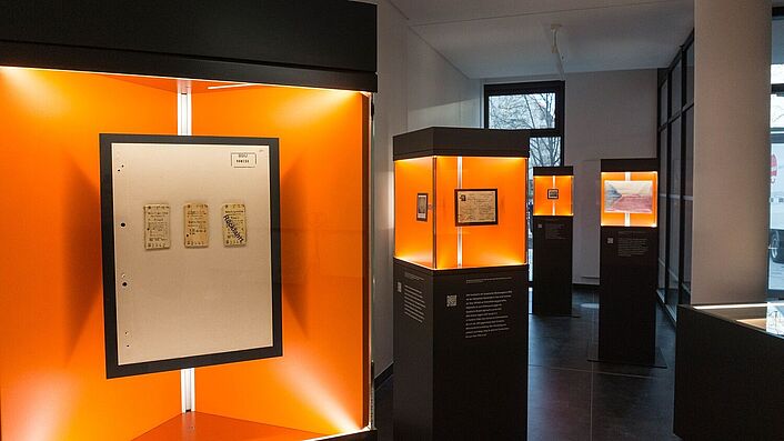 Blick in die Wanderausstellung. Zu sehen sind mehrere Stelen mit Glasvitrinen, in denen sich Dokumente, Fotos und Objekte befinden.