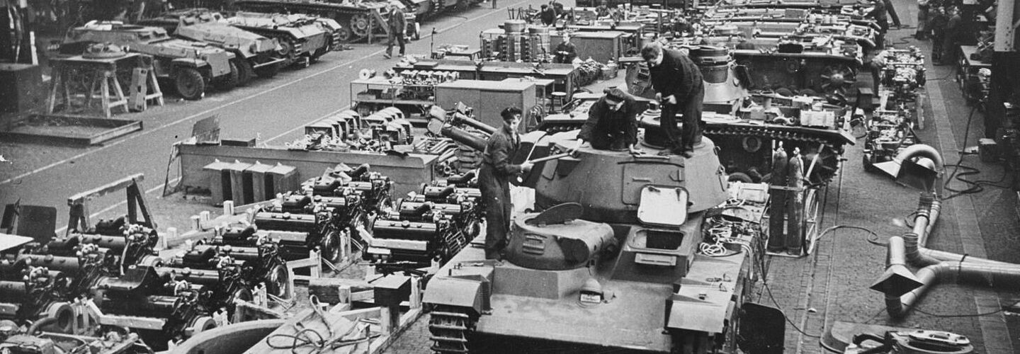Mehrere Panzer, Fahrzeuge und Fahrzeugteile in einem Rüstungswerk. Im Vordergrund ist ein "Neubaufahrzeug" (PzKpfw NbFz V, Rheinmetall) zu sehen.