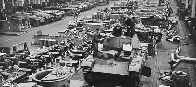 Mehrere Panzer, Fahrzeuge und Fahrzeugteile in einem Rüstungswerk. Im Vordergrund ist ein 'Neubaufahrzeug' (PzKpfw NbFz V, Rheinmetall) zu sehen.