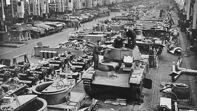 Mehrere Panzer, Fahrzeuge und Fahrzeugteile in einem Rüstungswerk. Im Vordergrund ist ein 'Neubaufahrzeug' (PzKpfw NbFz V, Rheinmetall) zu sehen.