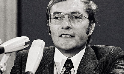 Auf dem Schwarz-Weiß-Foto ist der Gründer von „Der Spiegel“, Rudolf Augstein, auf einem FDP-Parteitag 1972 in seiner Funktion als Delegierter des Landesverbandes Nordrhein-Westfalen zu sehen. Vor Augstein stehen drei Mikrofone. Er selbst trägt eine Brille mit Metallrahmen, einen dunklen Anzug mit Karomuster und eine schwarze Krawatte mit bunten Mustern.