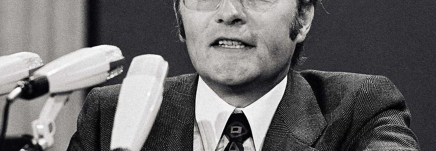 Rudolf Augstein auf dem FDP-Parteitag am 23. Oktober 1972 Auf dem Schwarz-Weiß-Foto ist der Gründer von „Der Spiegel“, Rudolf Augstein, auf einem FDP-Parteitag 1972 in seiner Funktion als Delegierter des Landesverbandes Nordrhein-Westfalen zu sehen. Vor Augstein stehen drei Mikrofone. Er selbst trägt eine Brille mit Metallrahmen, einen dunklen Anzug mit Karomuster und eine schwarze Krawatte mit bunten Mustern.
