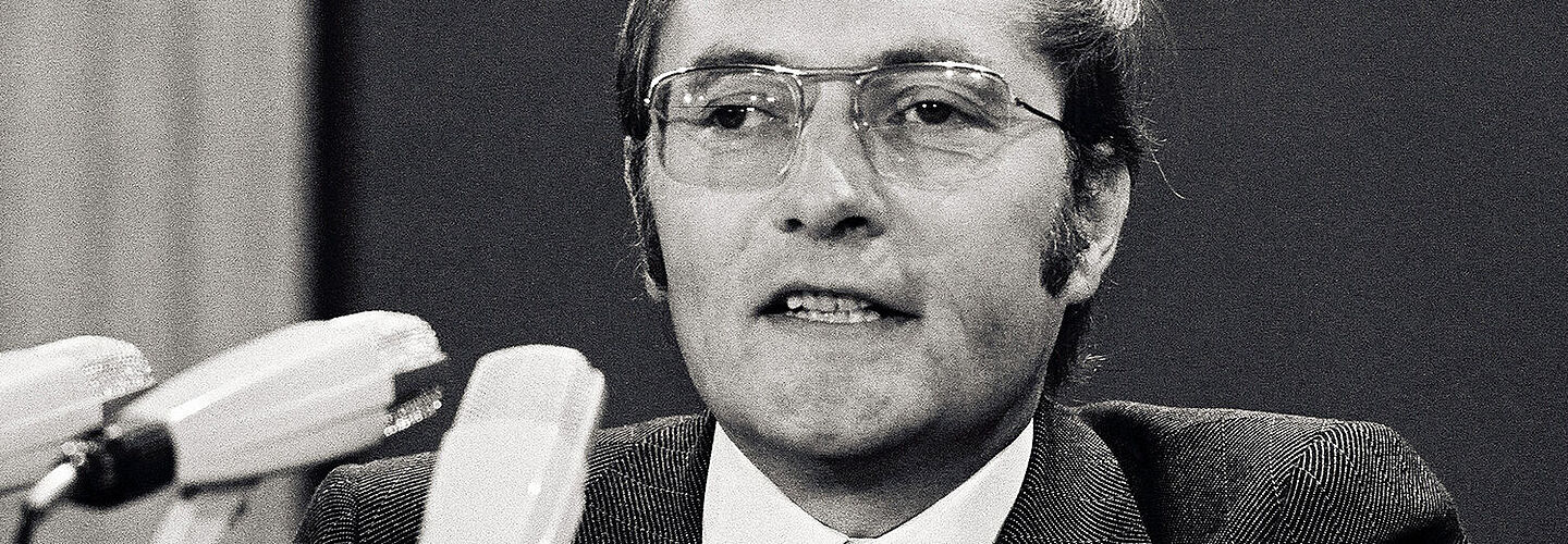 Auf dem Schwarz-Weiß-Foto ist der Gründer von „Der Spiegel“, Rudolf Augstein, auf einem FDP-Parteitag 1972 in seiner Funktion als Delegierter des Landesverbandes Nordrhein-Westfalen zu sehen. Vor Augstein stehen drei Mikrofone. Er selbst trägt eine Brille mit Metallrahmen, einen dunklen Anzug mit Karomuster und eine schwarze Krawatte mit bunten Mustern.