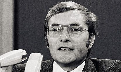 Auf dem Schwarz-Weiß-Foto ist der Gründer von 'Der Spiegel', Rudolf Augstein, auf einem FDP-Parteitag 1972 in seiner Funktion als Delegierter des Landesverbandes Nordrhein-Westfalen zu sehen. Vor Augstein stehen drei Mikrofone. Er selbst trägt eine Brille mit Metallrahmen, einen dunklen Anzug mit Karomuster und eine schwarze Krawatte mit bunten Mustern.