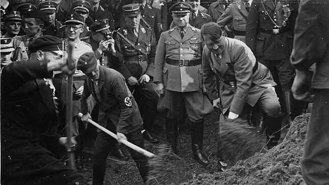 Adolf Hitler und ein weiterer Mann mit Hakenkreuz-Emblem auf dem Jackenärmel schippen mit Schaufeln Sand auf eine Haufen. Zahlreiche uniformierte Männer schauen zu.