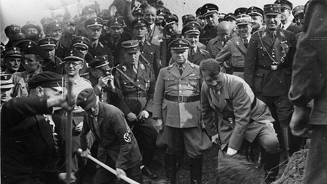 Adolf Hitler und ein weiterer Mann mit Hakenkreuz-Emblem auf dem Jackenärmel schippen mit Schaufeln Sand auf eine Haufen. Zahlreiche uniformierte Männer schauen zu.