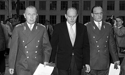 Die Generale Hans Speidel und Adolf Heusinger in Uniform halten ihre Ernennungsurkunden in den Händen. Zwischen ihnen geht Verteidigungsminister Theodor Blank im Anzug. Im Hintergrund sind Presseteams zu erkennen.