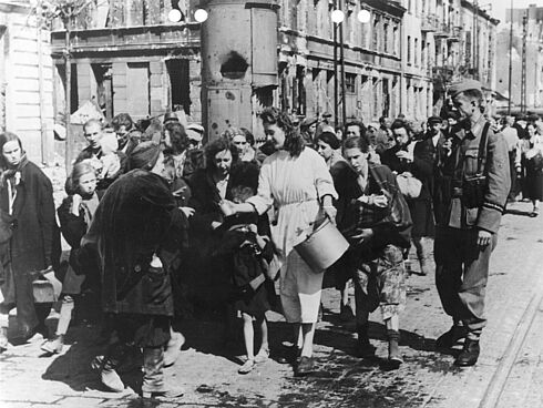 Zivilistinnen und Zivilisten laufen, von deutschen Soldaten begleitet, eine Straße entlang. Eine Frau in weißem Kleid verteilt Lebensmittel aus einem Eimer an sie.