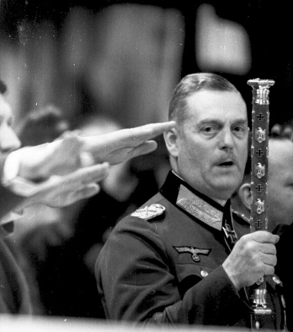 Wilhelm Keitel hält einen Marschallstab in die Luft. Neben ihm zeigen Personen den Hitlergruß.