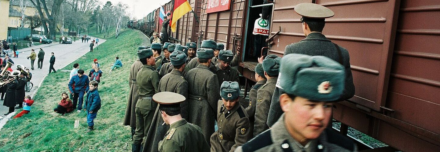 Soldaten der 47. Panzerdivison der GUS-Streitkräfte drängen sich vor einem offenen Güterwagen. Auf dem Bahndamm sitzen Zivilistinnen und Zivilisten, im Hintergrund spielt eine Kapelle.