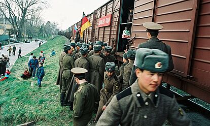 Soldaten der 47. Panzerdivison der GUS-Streitkräfte drängen sich vor einem offenen Güterwagen. Auf dem Bahndamm sitzen Zivilistinnen und Zivilisten, im Hintergrund spielt eine Kapelle.