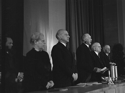 Schwarz-Weiß-Aufnahme mit fünf Herren und einer Frau in formeller Kleidung, hinter einem langen Holztisch stehend